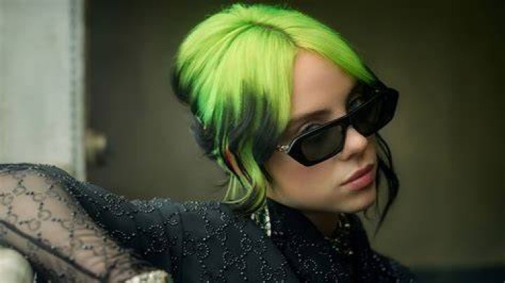 Billie Eilish Wearing a green top with jewelry in her ears and neck and round black spectacles.